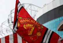 Red Manchester United flag with club crest blows in the wind near Old Trafford stadium.